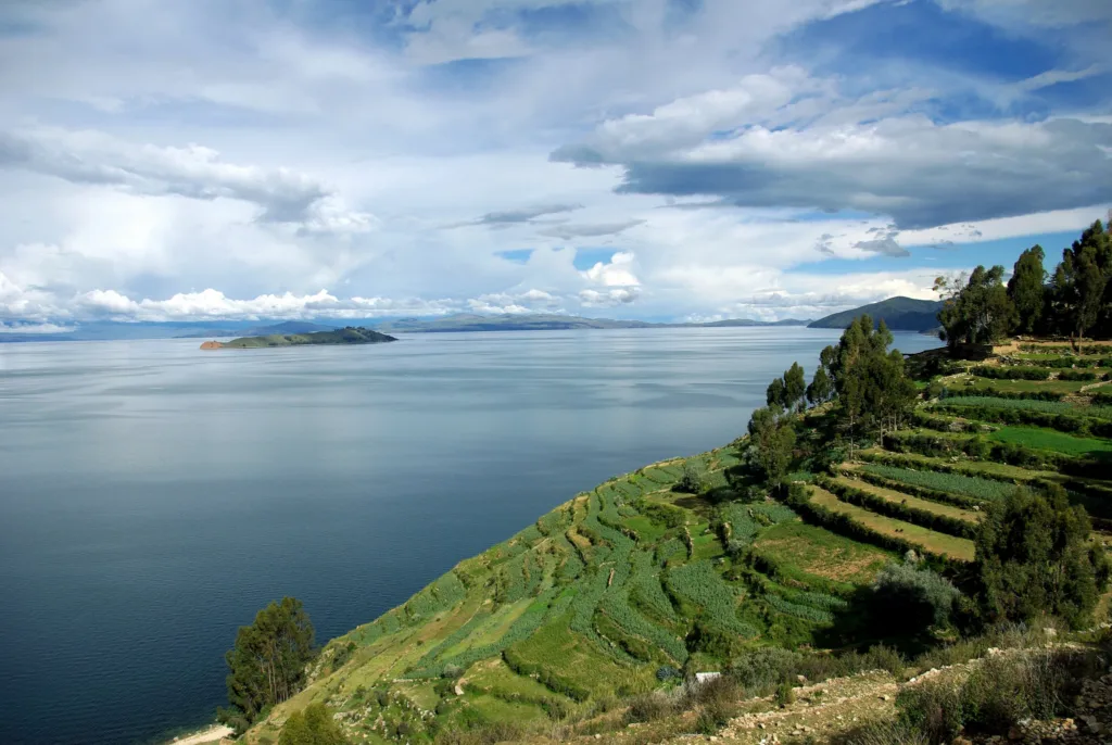Zelené zemědělské terasy na svahu nad klidnou hladinou jezera Titicaca pod zataženou oblohou.