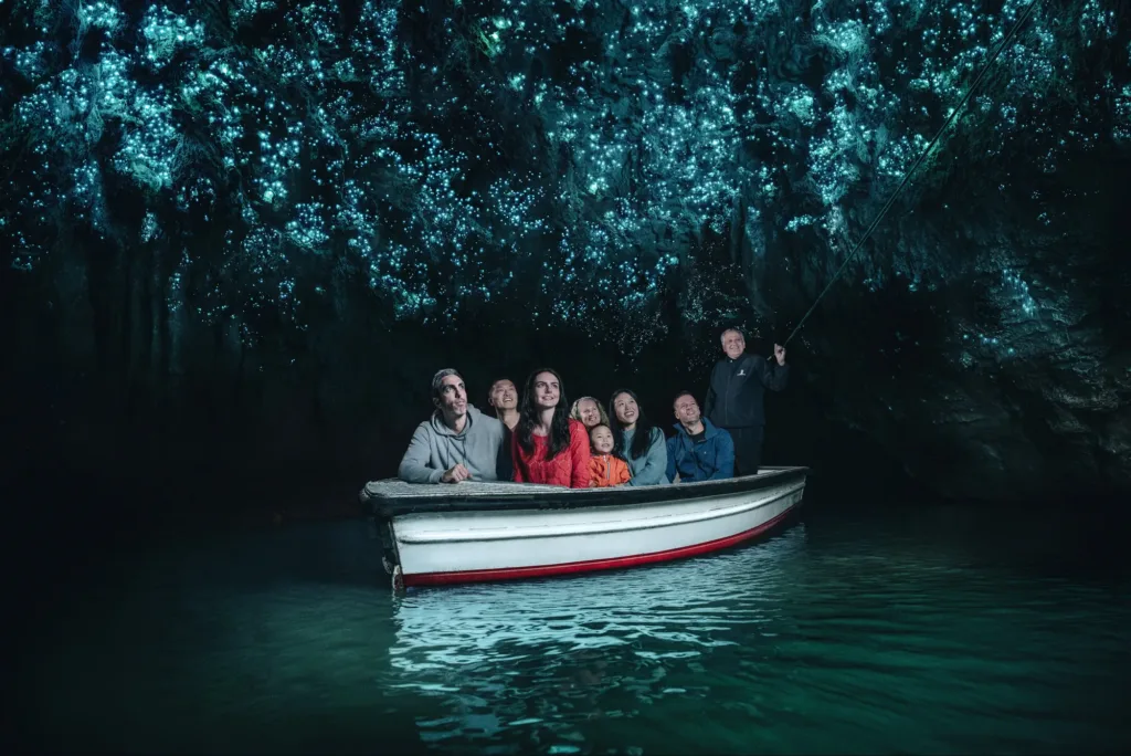 Skupina turistů na lodi projíždí podzemní jeskyní, jejíž strop září tisíci modrozelenými světélky bioluminiscenčních larev
