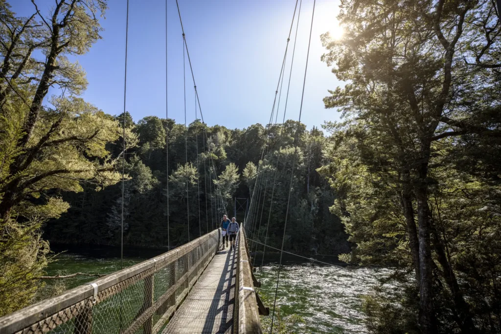 Dva turisté s batohy přecházejí dřevěný visutý most nad zelenou řekou obklopenou hustým lesem