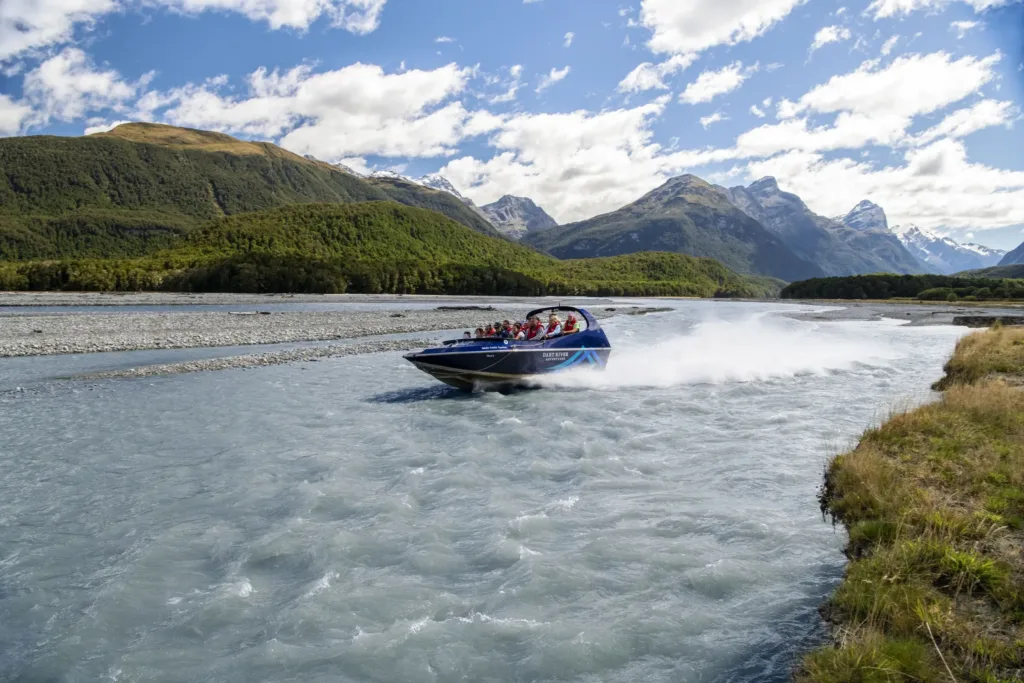 Modrý jet boat s cestujícími v záchranných vestách projíždí rychlostí po řece obklopené horami a lesnatými kopci