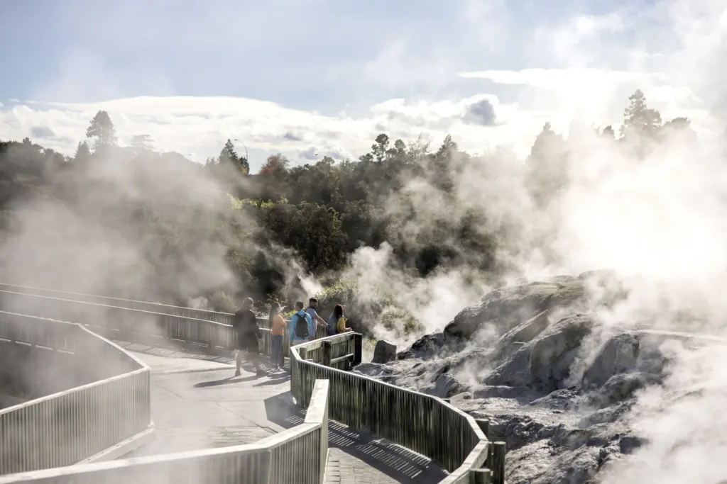 Dřevěná vyhlídková stezka s turisty procházející geotermální oblastí, kde ze skalnatého terénu stoupá hustá pára