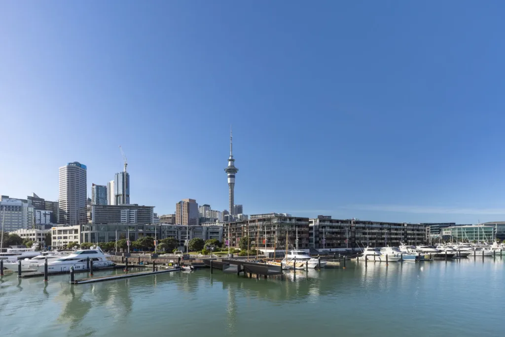 Pohled na siluetu města Auckland s vysokou vysílací věží Sky Tower, mrakodrapy a přístavní marínou plnou jachet za jasného dne