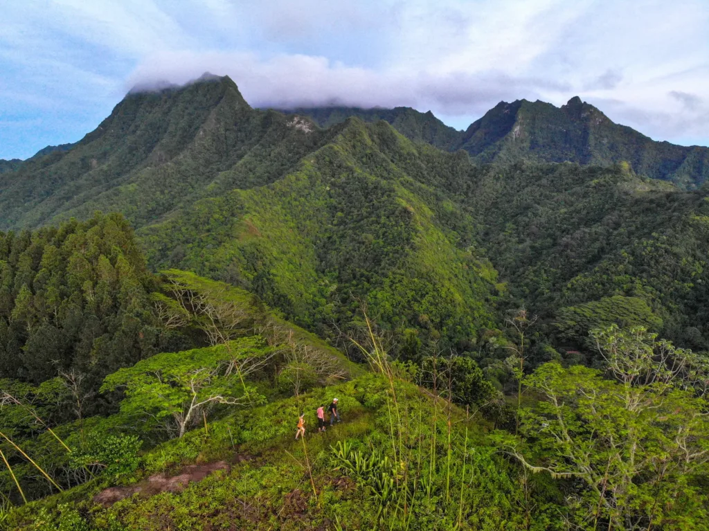 Turisté na stezce mezi tropickou vegetací s výhledem na zelené sopečné hory zahalené mraky.
