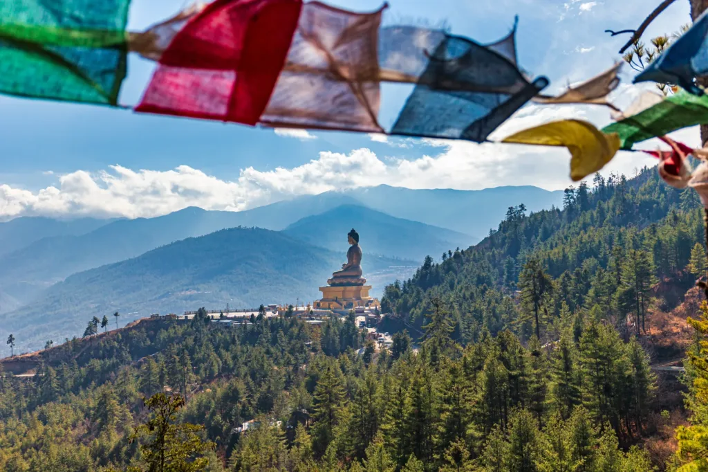 Barevné modlitební praporky nad pohledem na sochu Buddha Dordenma a zalesněné himálajské svahy.