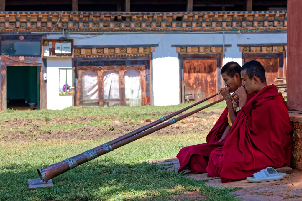 Dva buddhističtí mniši v červených rouchách hrají na dlouhé tradiční rohy před zdobenou budovou.