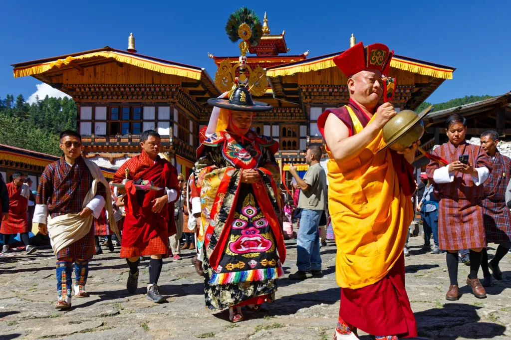 Mniši a účastníci v tradičních oděvech při buddhistickém festivalu před zdobenou klášterní budovou v Bhútánu.
