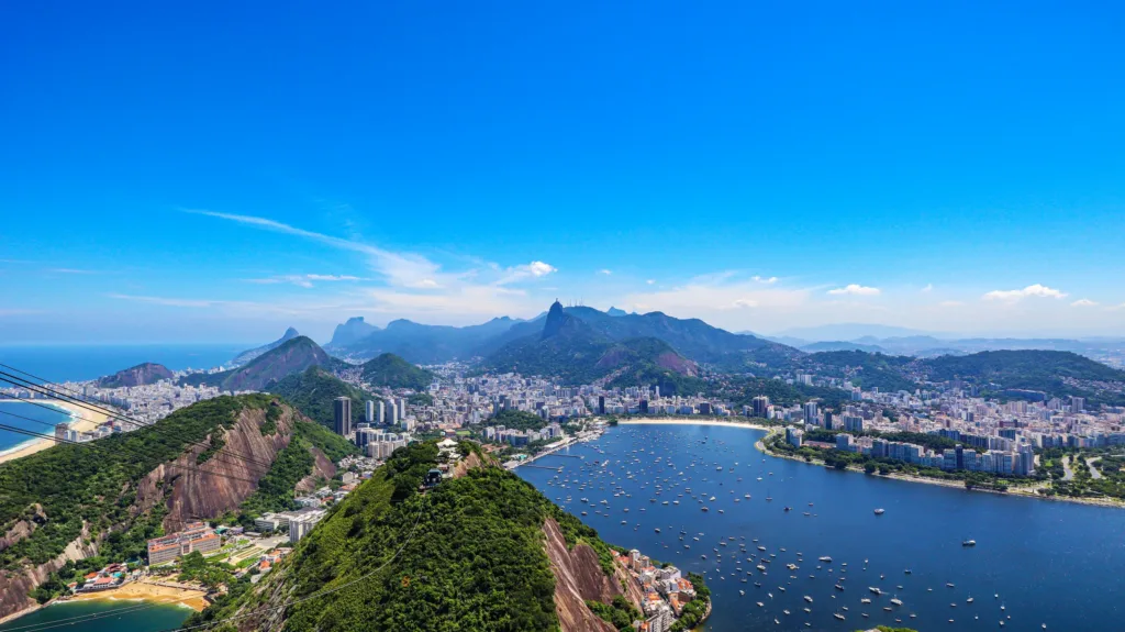 Pohled na Rio de Janeiro se zátokou Guanabara, zelenými kopci, městskou zástavbou a loděmi na hladině.