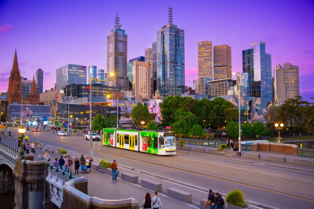 Večerní pohled na centrum Melbourne s mrakodrapy, zelenou tramvají na mostě a fialovým nebem po západu slunce