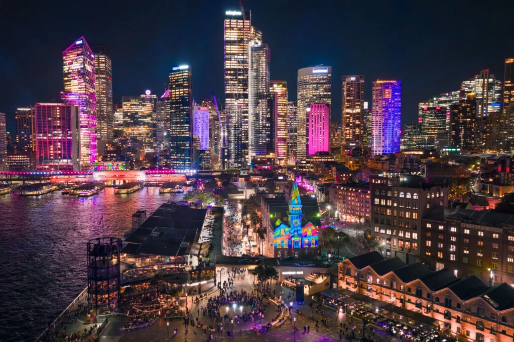 Noční pohled na mrakodrapy Sydney nasvícené barevnými světly, s Circular Quay a nábřežím plným lidí u vody