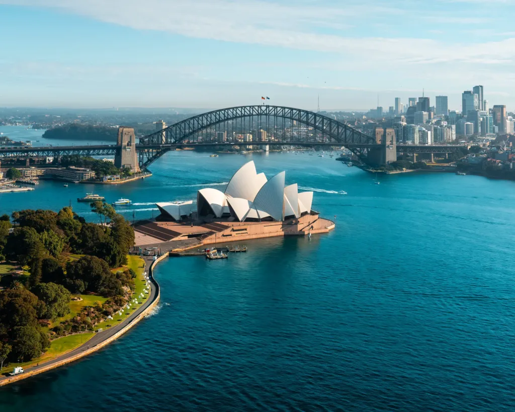 Letecký pohled na Sydney Harbour s bílou budovou opery ve tvaru mušlí, ocelovým obloukovým mostem a mrakodrapy centra města v pozadí