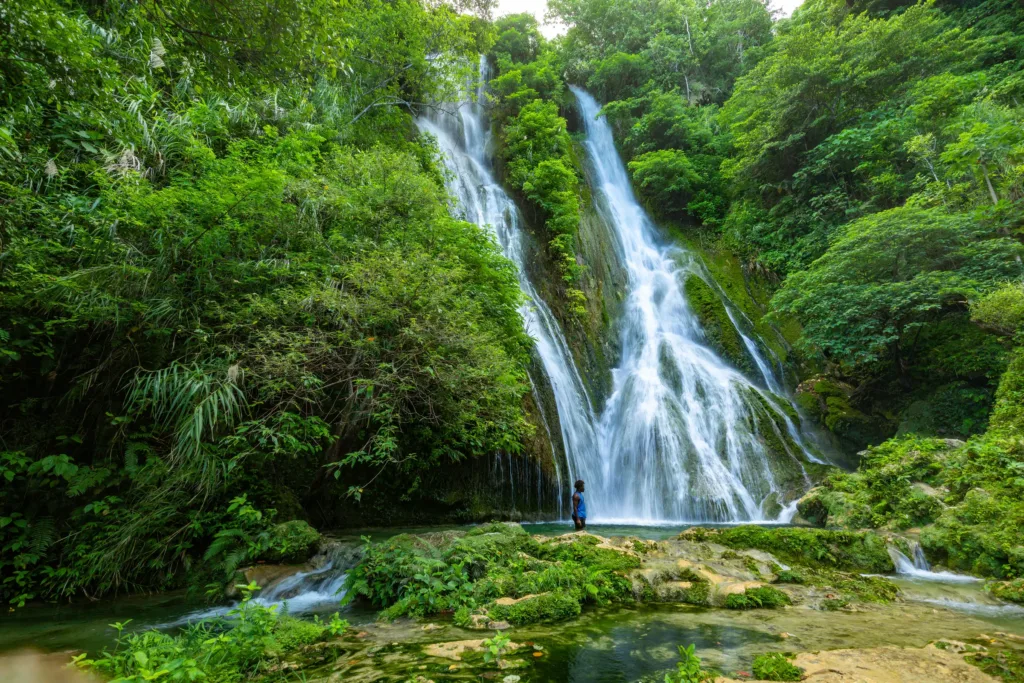 Vysoký vodopád stékající po skalách obklopených hustou tropickou zelení, u paty stojí člověk.
