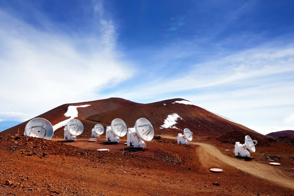 Řada bílých radioteleskopů na kamenité sopečné krajině pod červenohnědým kuželem Mauna Kea.