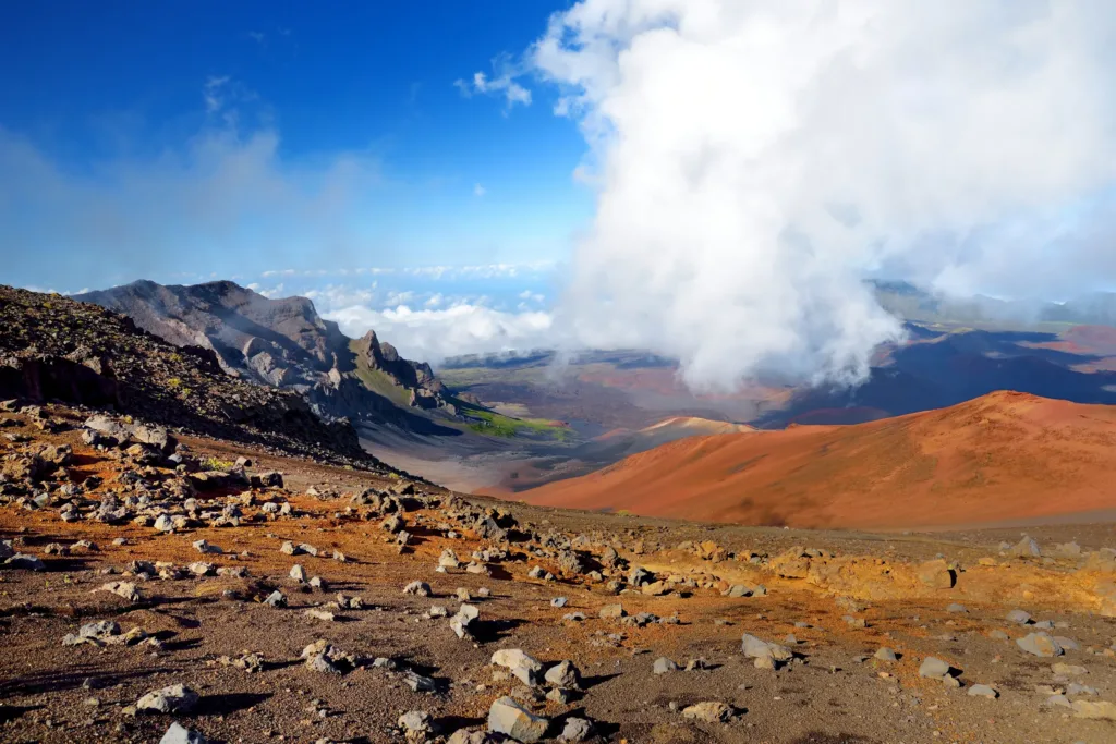 Pohled z kamenitého sopečného svahu na červenohnědé kužely a mraky nad kráterem Haleakalā.