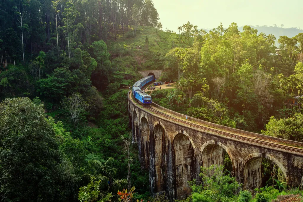 Modrý vlak přejíždí kamenný Most Devíti oblouků obklopený tropickou zelení na Srí Lance.