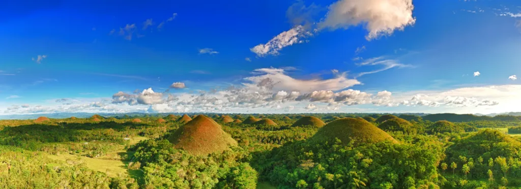Zelené kuželovité kopce Chocolate Hills obklopené tropickou vegetací pod modrou oblohou s mraky.