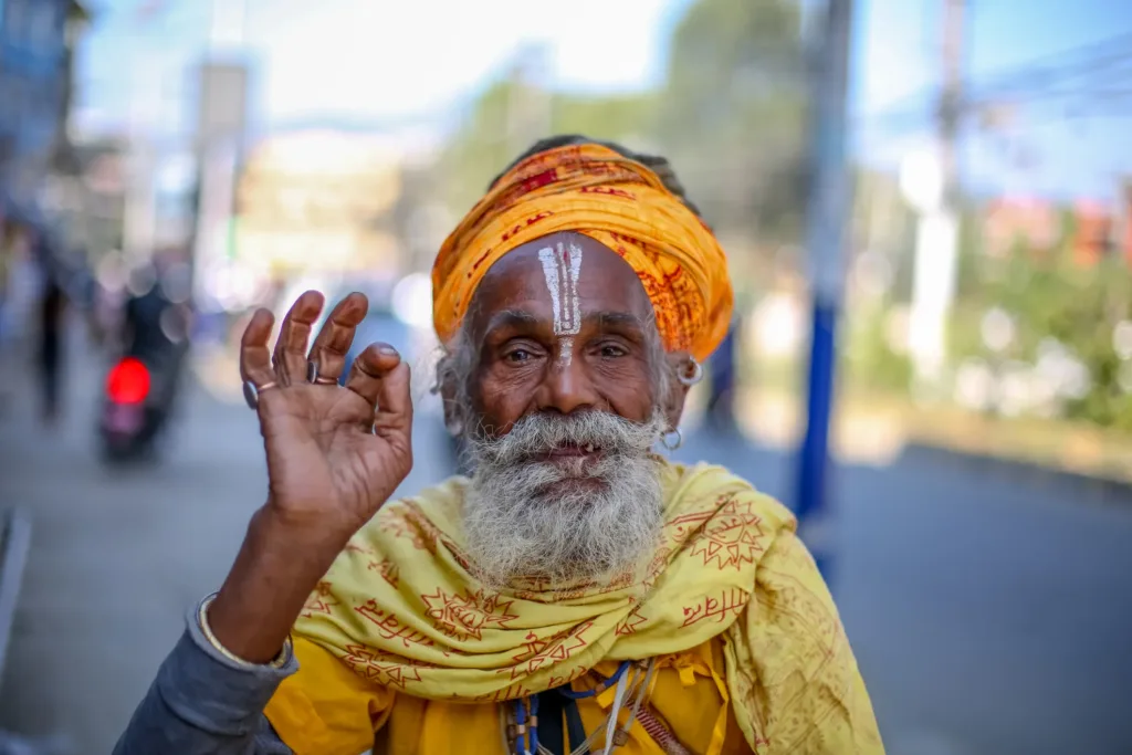 Usmívající se hinduistický asketa se žlutým turbanem a pomalovaným čelem gestikuluje do fotoaparátu.
