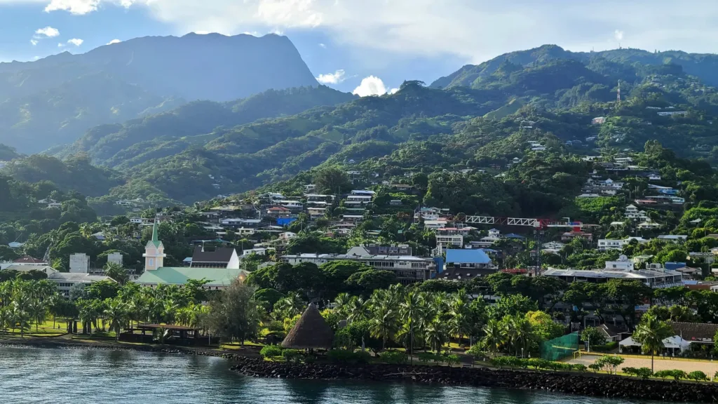 Pobřežní vesnice na ostrově Tahiti s bílým kostelem, tropickou vegetací a zelenými horami v pozadí.