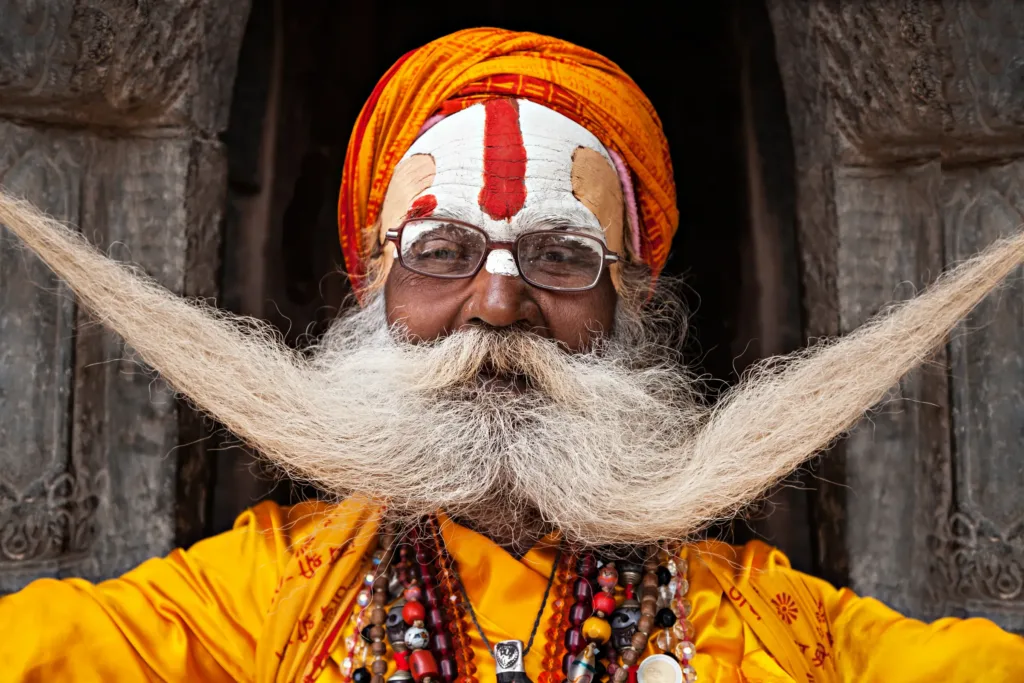 Usměvavý hinduistický sádhu v oranžovém turbanu s dlouhým roztaženým knírem a pomalovaným čelem.