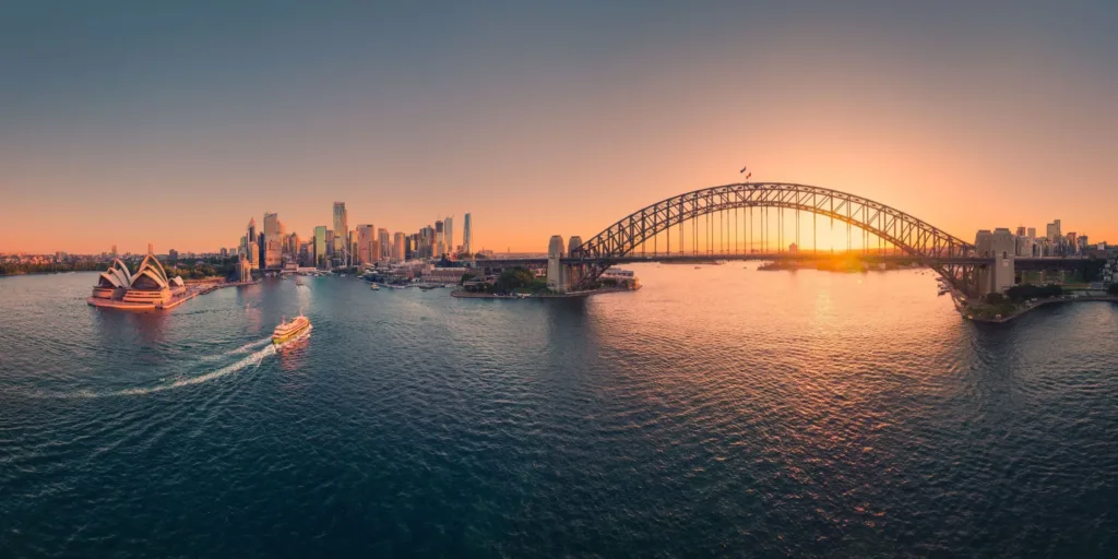 Pohled na přístav v Sydney se Sydney Opera House, trajekty a mostem Sydney Harbour Bridge při západu slunce.