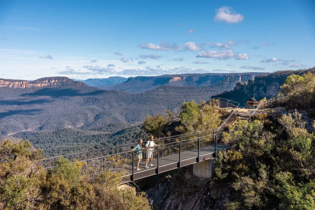 Turisté na kovové vyhlídkové lávce nad zalesněným údolím v pohoří Blue Mountains.