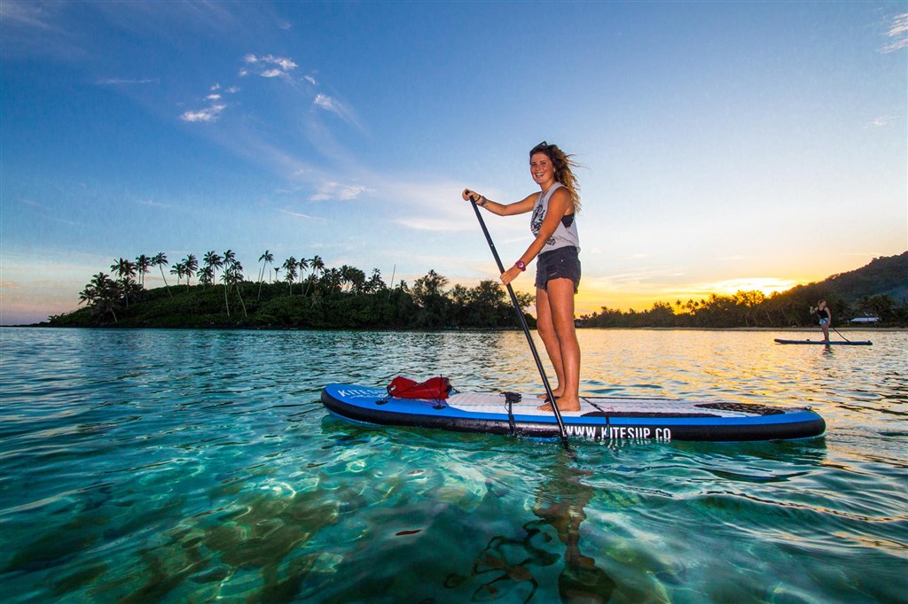Žena na paddleboardu projíždí průzračnou tyrkysovou lagunou s palmovým ostrůvkem v pozadí při východu slunce