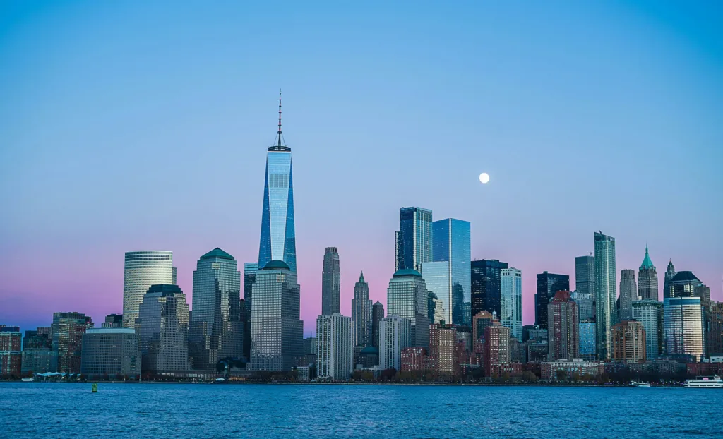 Mrakodrapy Dolního Manhattanu s dominantní věží One World Trade Center při západu slunce, v pozadí vycházející úplněk, v popředí hladina řeky