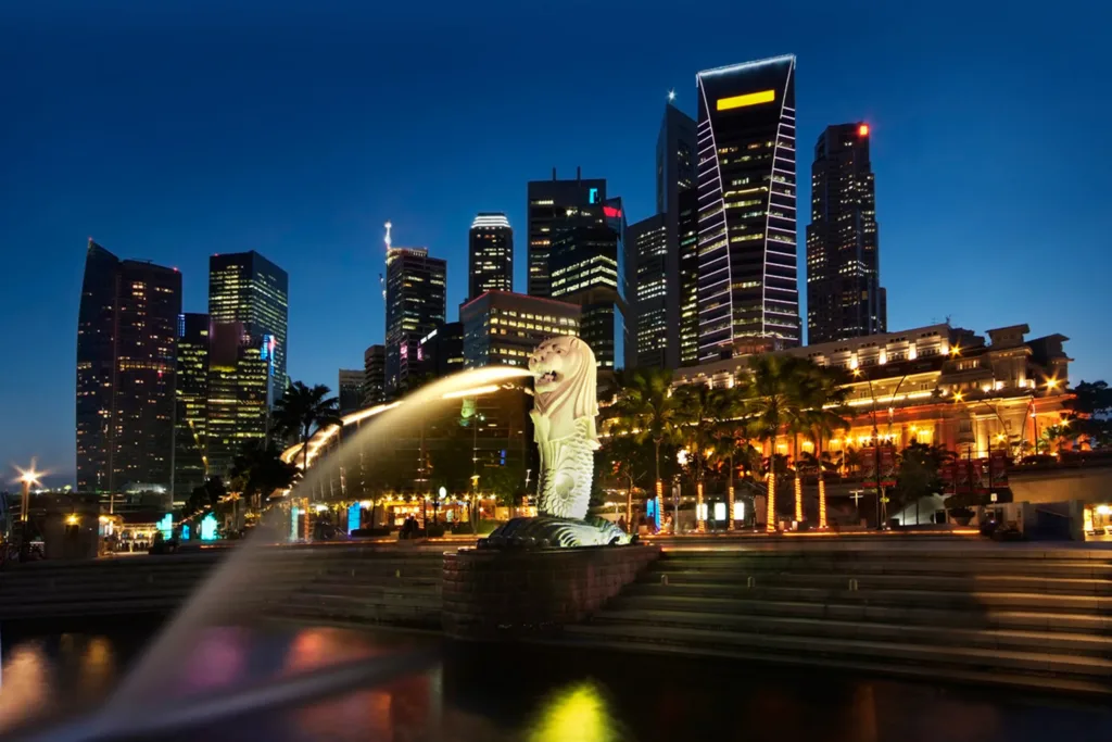 Osvětlená socha Merlion chrlící vodu do zálivu, v pozadí mrakodrapy nasvíceného nočního Singapuru